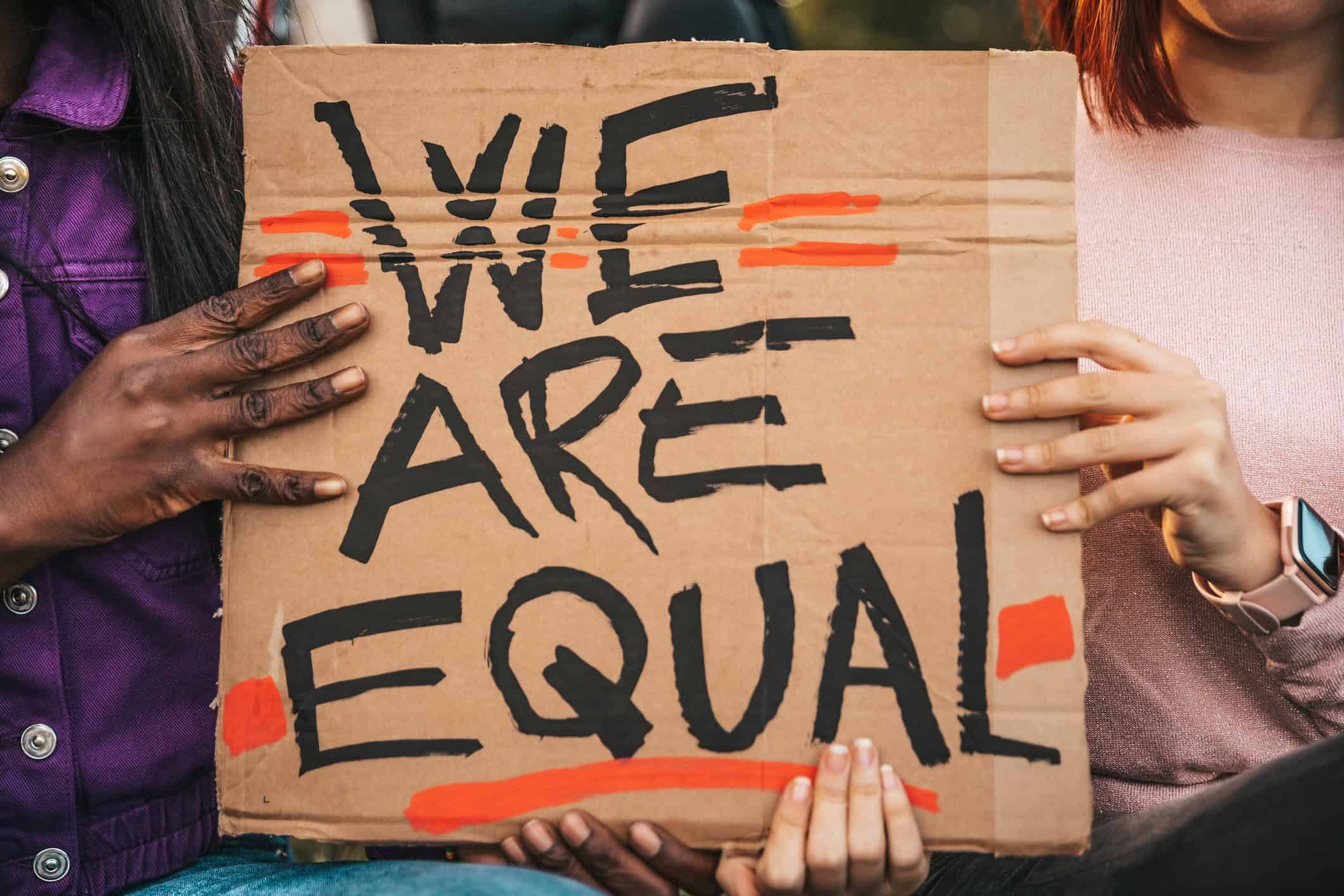 Multiple hands holding cardboard sign reading "WE ARE EQUAL" with orange highlighting