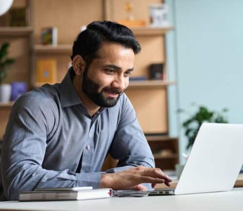 Smiling man in gray shirt working on laptop at home office desk