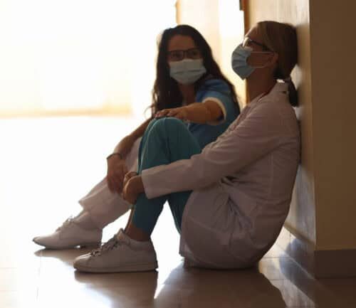 Two exhausted female doctors in masks sitting on hospital corridor floor