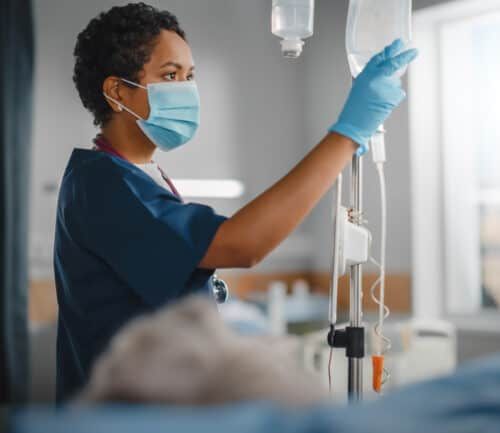 Black nurse in mask adjusting IV drip in hospital patient room