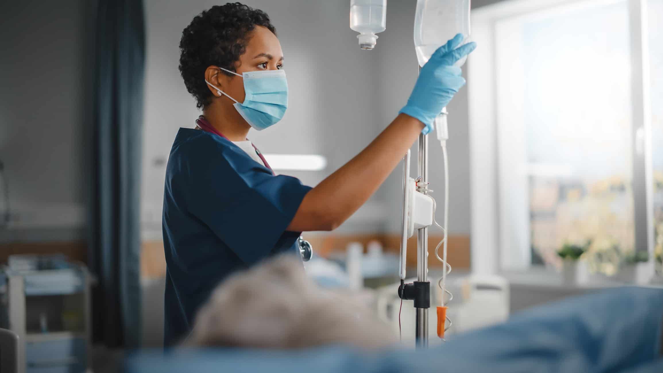 Black nurse in mask adjusting IV drip in hospital patient room