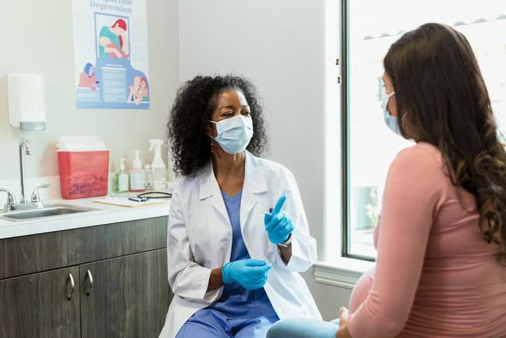 Doctor in mask gestures while consulting with pregnant patient in medical office