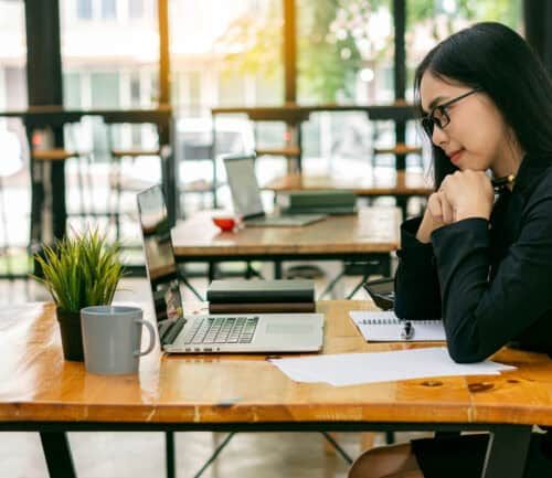 Asian woman in glasses working on laptop at wooden desk in bright office