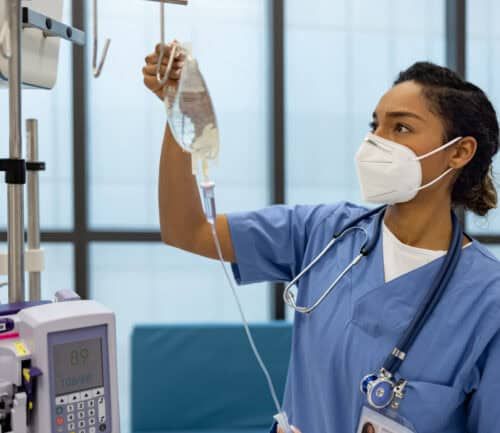 Nurse in blue scrubs and mask preparing IV drip bag in hospital room