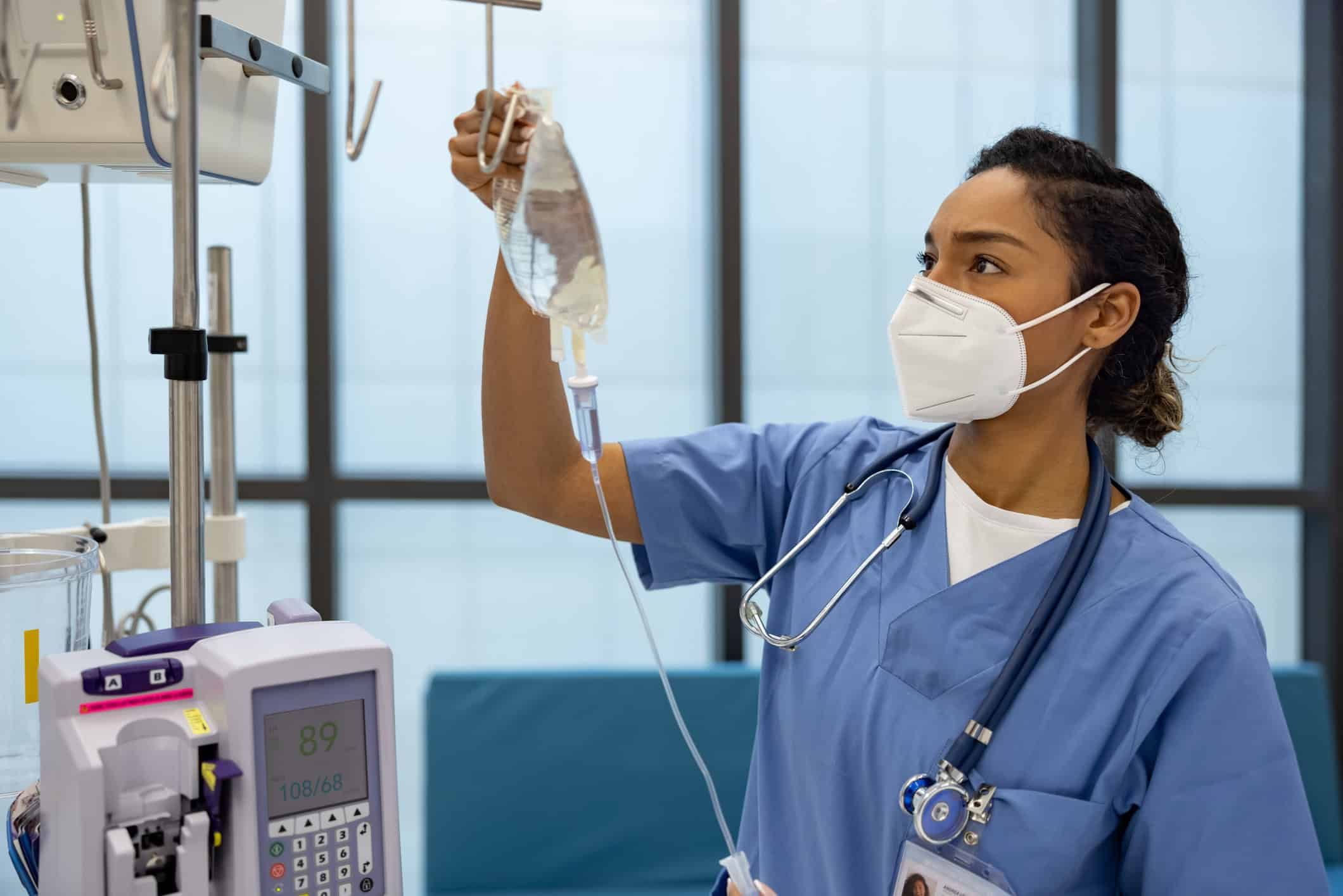 Nurse in blue scrubs and mask preparing IV drip bag in hospital room