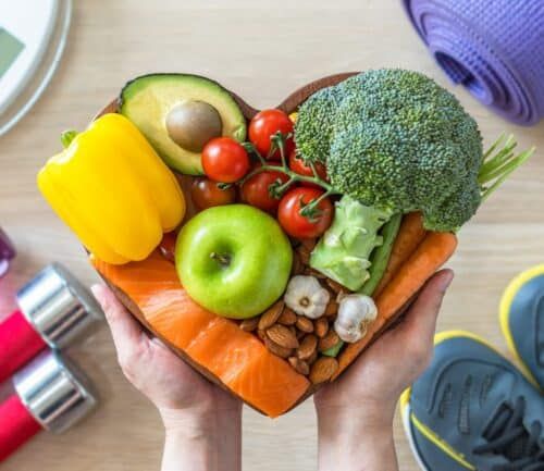 Hands holding heart-shaped bowl of healthy foods with exercise equipment nearby
