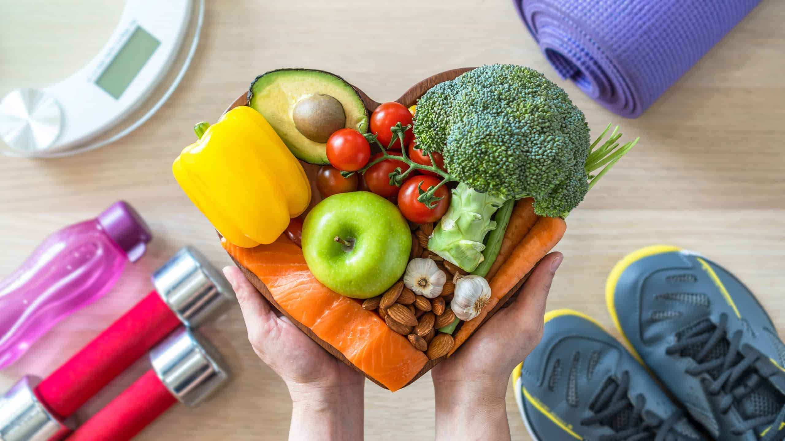 Hands holding heart-shaped bowl of healthy foods with exercise equipment nearby
