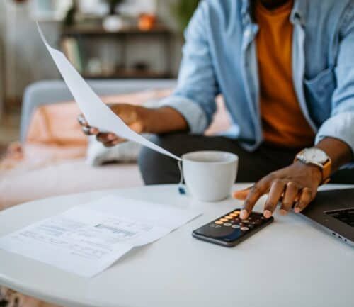 Person reviewing financial documents with calculator and laptop on kitchen table