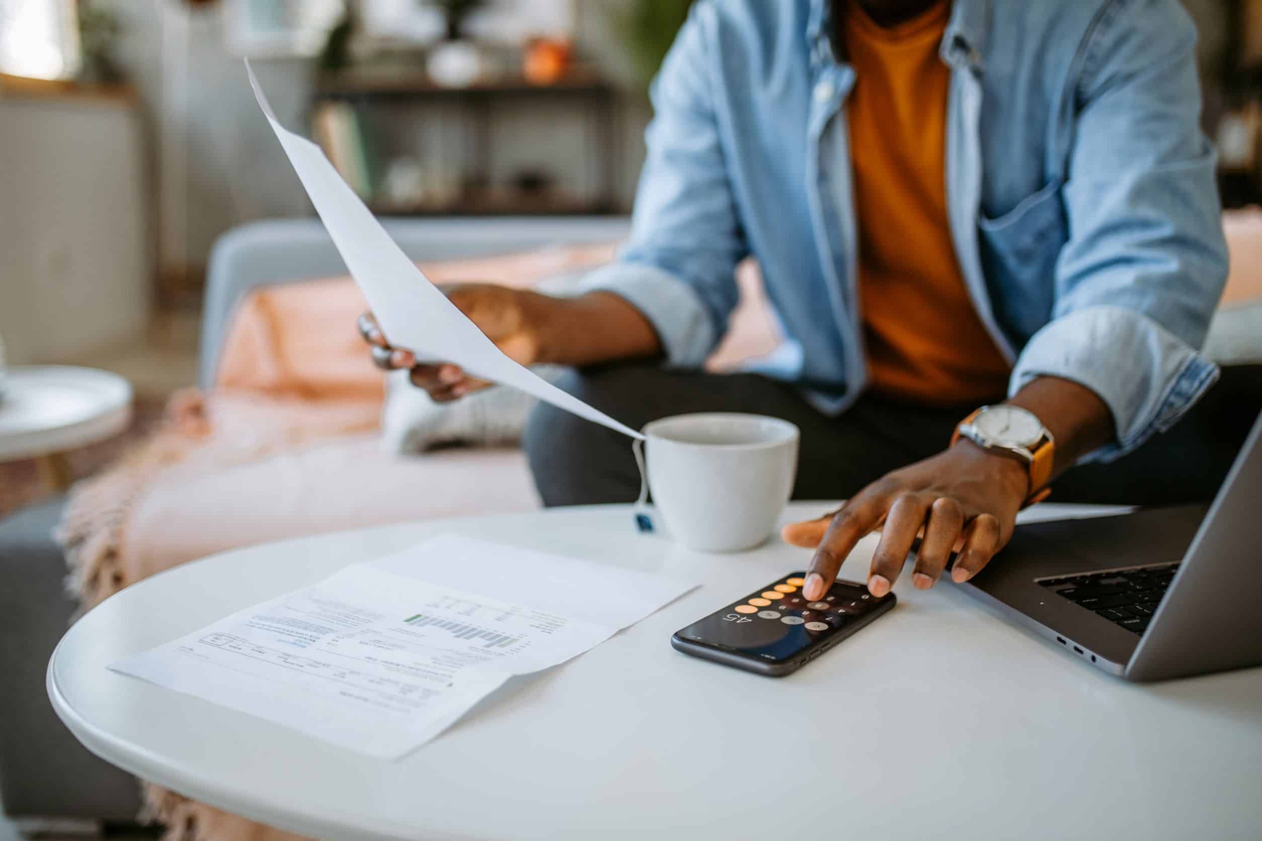 Person reviewing financial documents with calculator and laptop on kitchen table