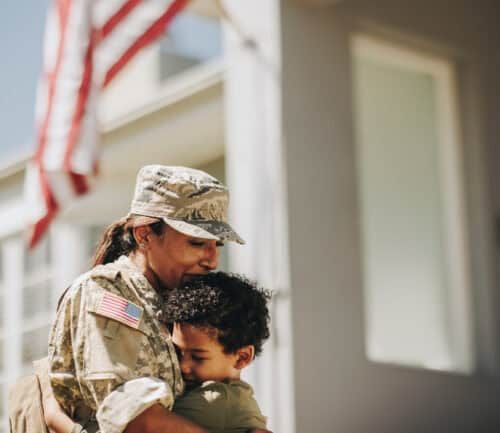 Female soldier in camouflage uniform embracing young boy with American flag in background