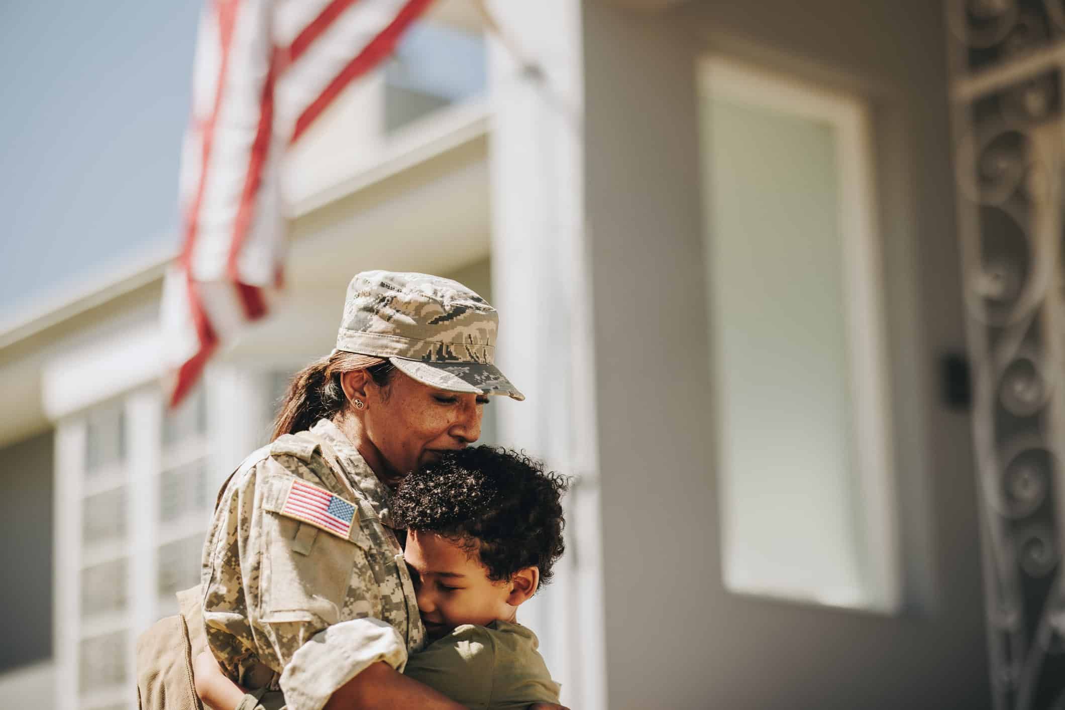 Female soldier in camouflage uniform embracing young boy with American flag in background