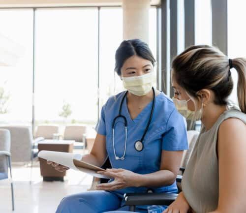 Female nurse in blue scrubs consulting with masked patient holding medical records