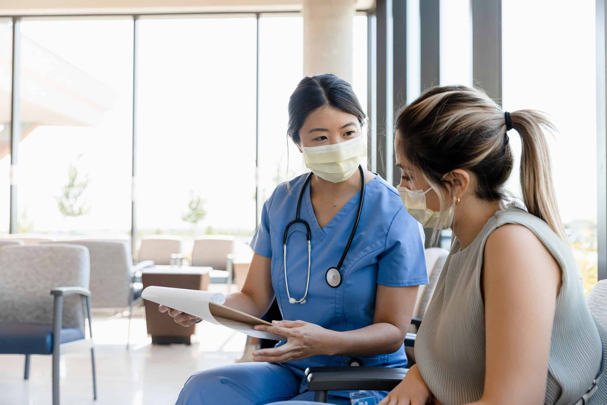 Female nurse in blue scrubs consulting with masked patient holding medical records