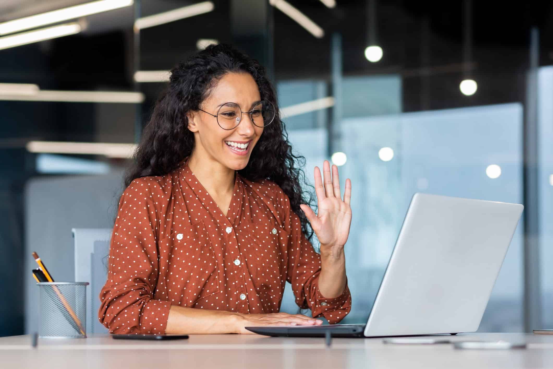 Hispanic businesswoman waving during video call on laptop in modern office