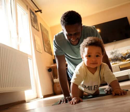 Father encouraging toddler to crawl on hardwood floor in bright living room