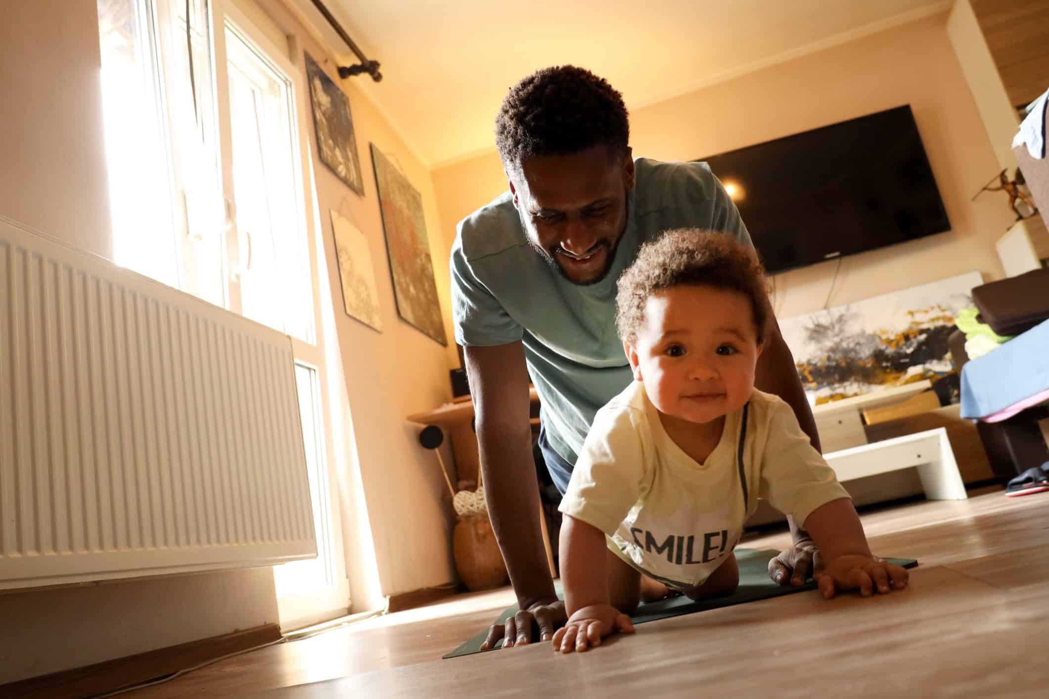 Father encouraging toddler to crawl on hardwood floor in bright living room