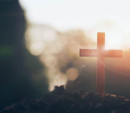 Wooden cross silhouetted against soft sunlight with blurred natural background.