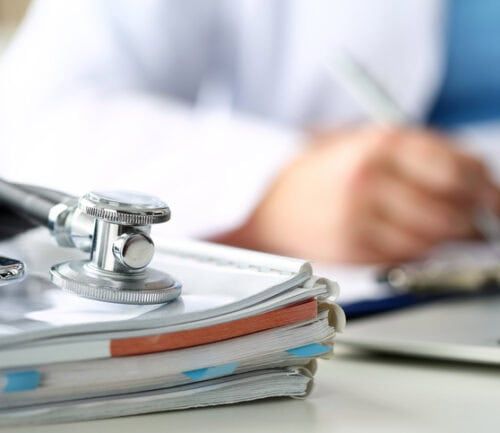 Stethoscope resting on stack of medical forms with doctor writing in background