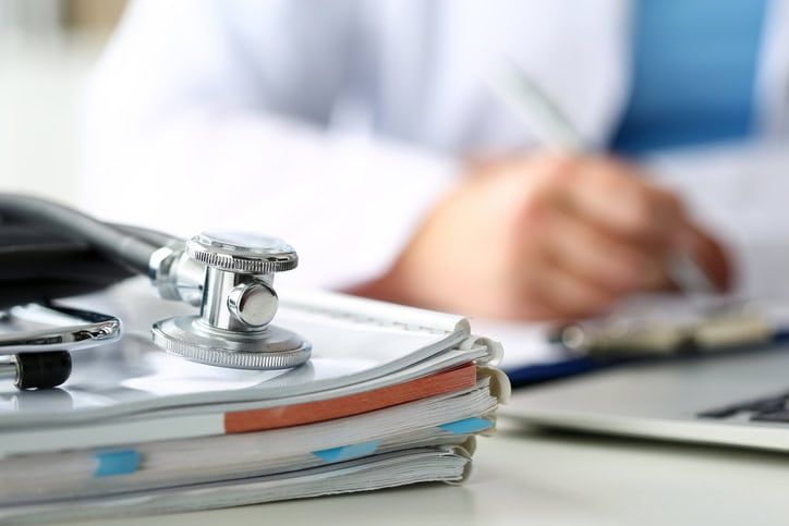Stethoscope resting on stack of medical forms with doctor writing in background