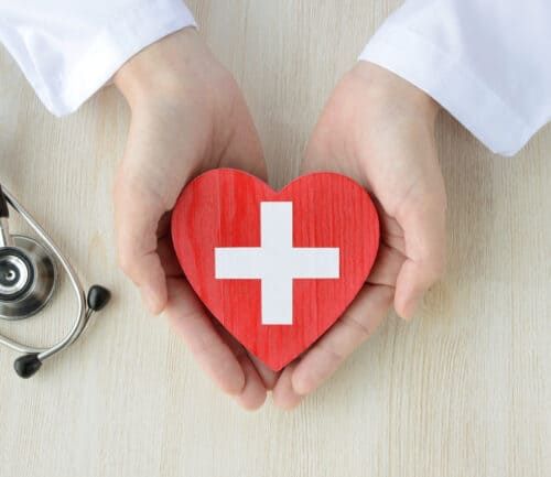 Hands in white coat holding red heart with medical cross, stethoscope nearby