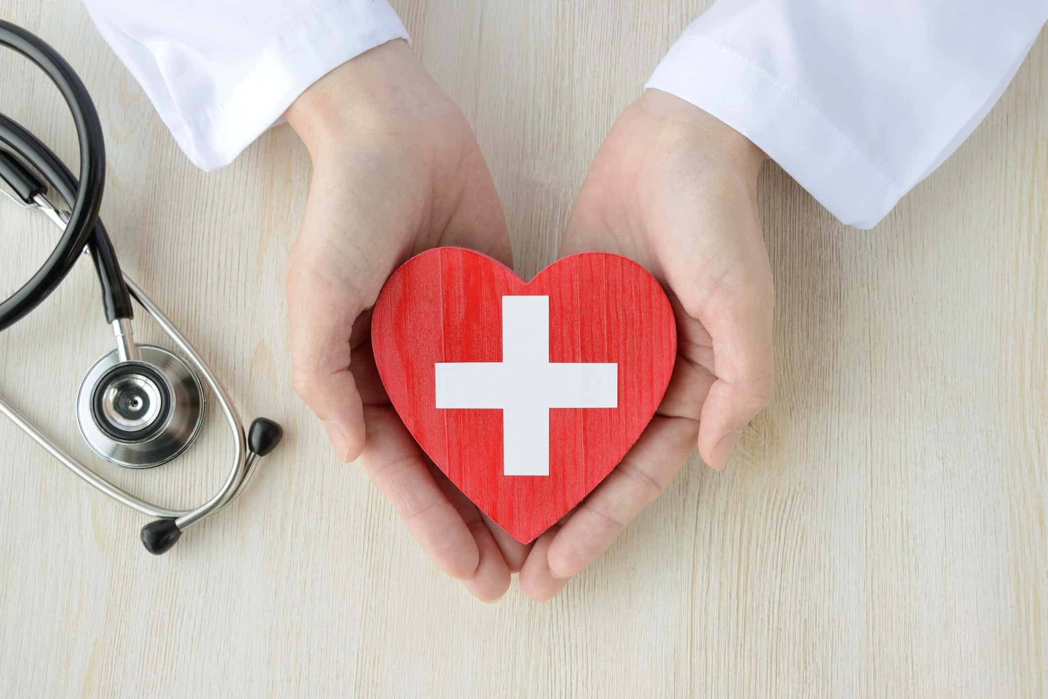 Hands in white coat holding red heart with medical cross, stethoscope nearby
