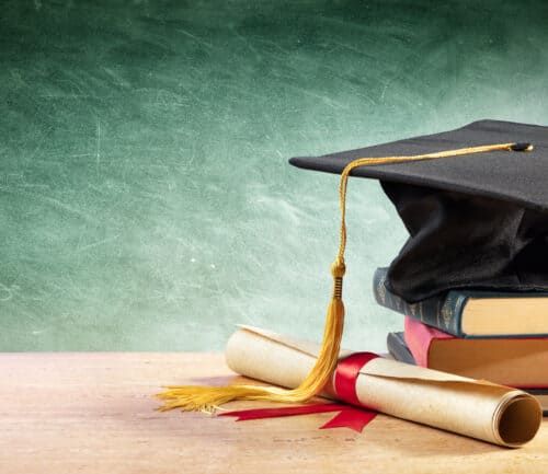 Graduation cap with gold tassel sits on stack of books beside rolled diploma