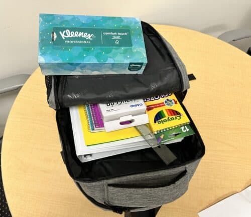 Open gray backpack containing school supplies, tissues, and crayons on wooden table