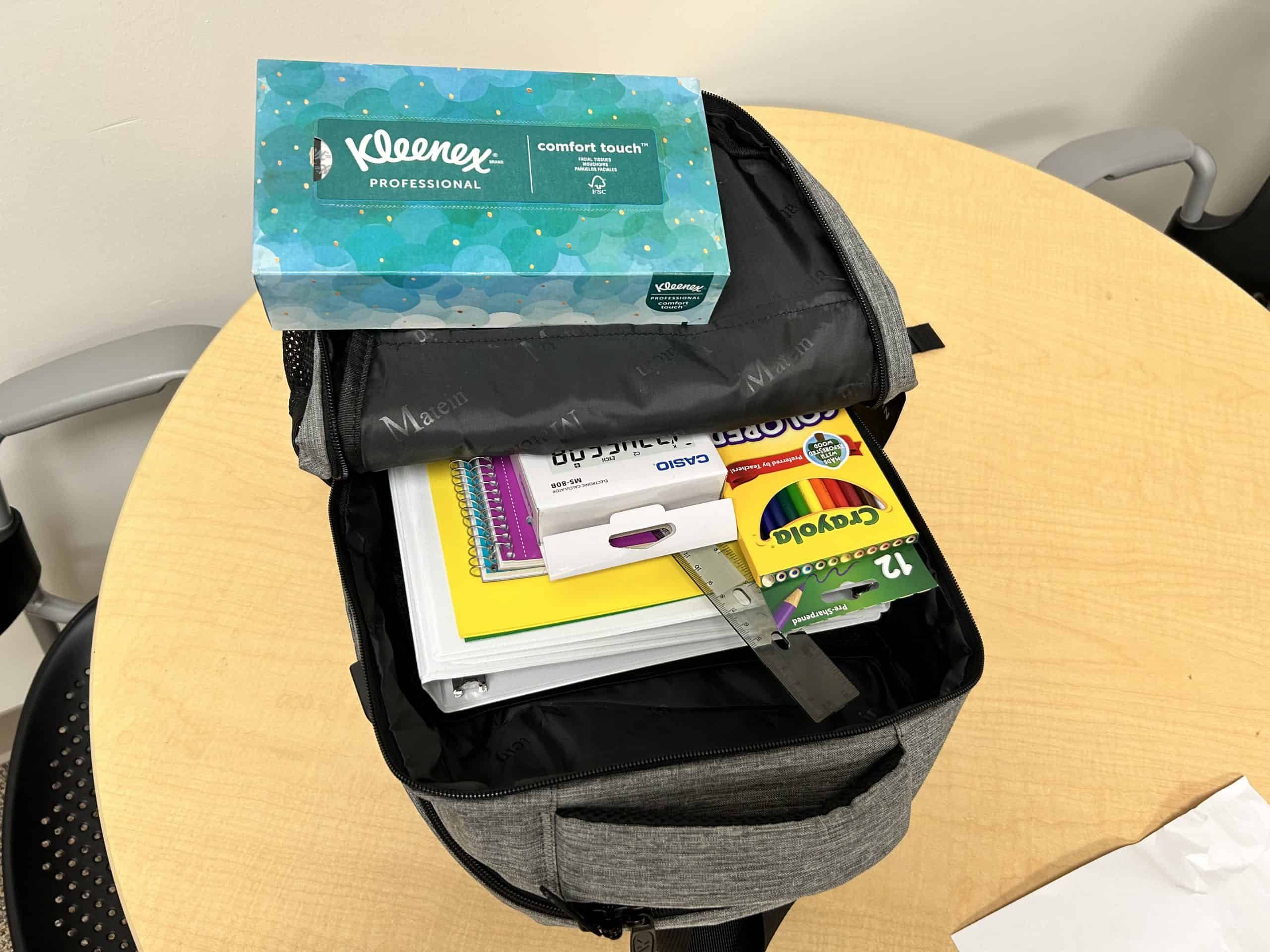 Open gray backpack containing school supplies, tissues, and crayons on wooden table