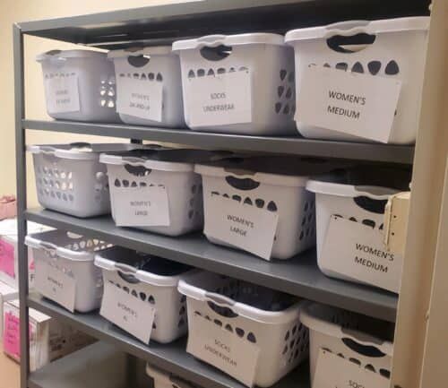 Organized shelving unit with labeled white laundry baskets for women's clothing donations by size.