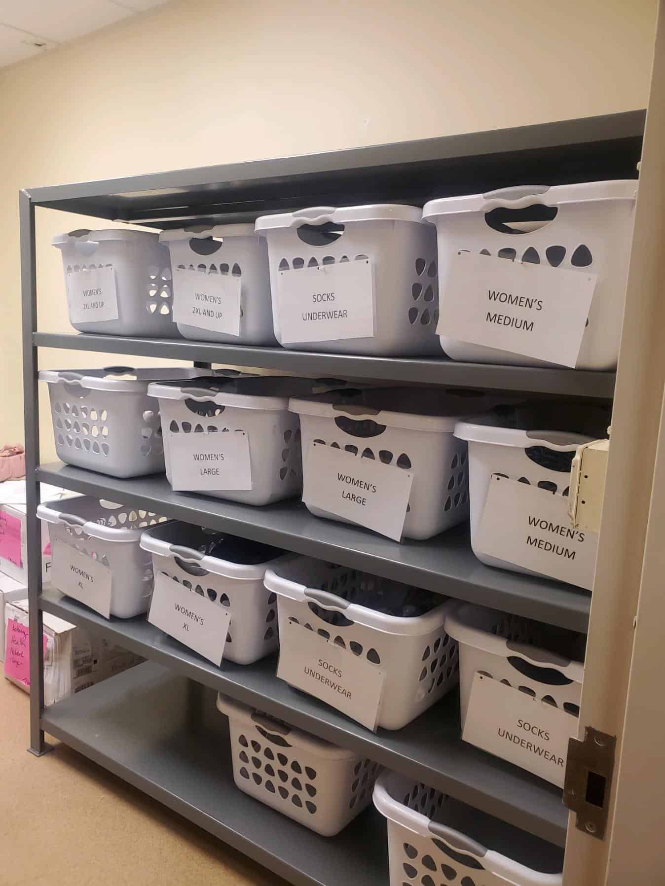Organized shelving unit with labeled white laundry baskets for women's clothing donations by size.