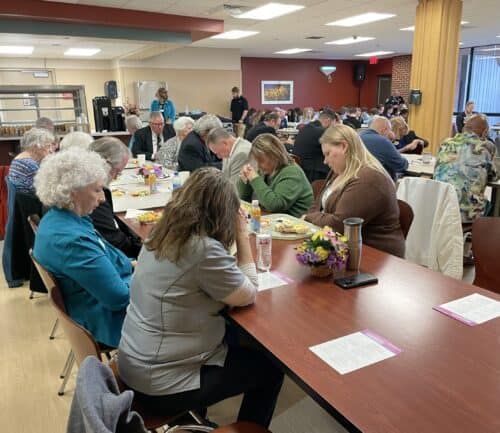 People gathered around tables eating meals in a community dining room setting.
