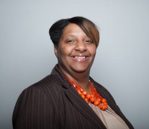 Professional headshot of Jennifer Woods wearing dark blazer with orange beaded necklace