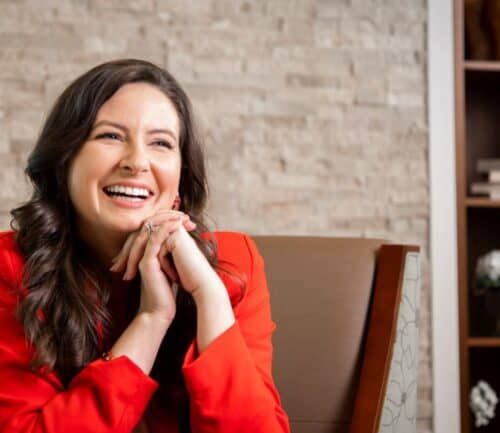 Woman in red blazer laughing with hands clasped under chin in office setting