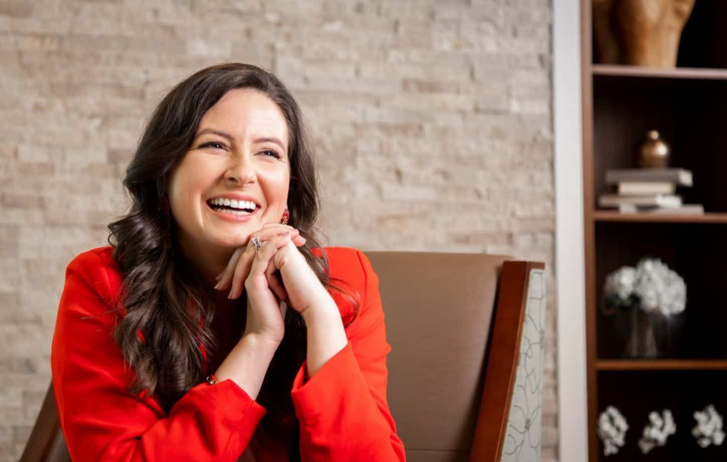 Woman in red blazer laughing with hands clasped under chin in office setting