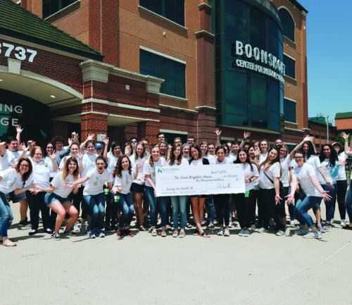 Large group of students in matching white shirts celebrating with oversized check outside college building