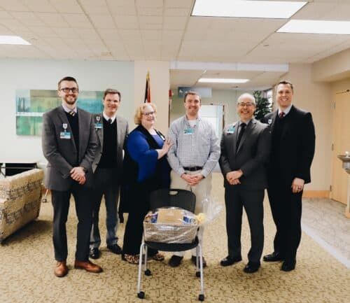 Six healthcare professionals in business attire posing together in hospital lobby with gift basket.