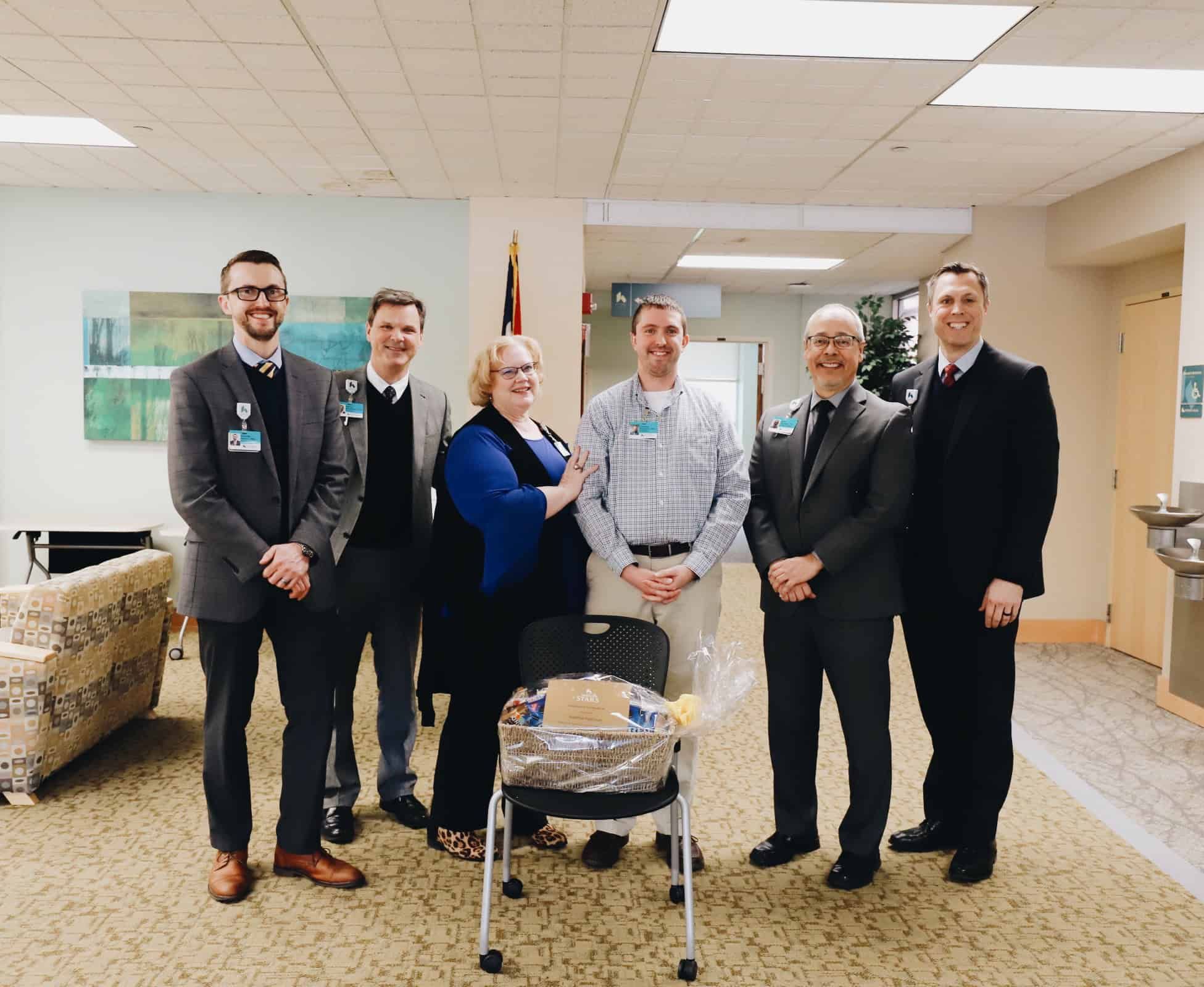 Six healthcare professionals in business attire posing together in hospital lobby with gift basket.
