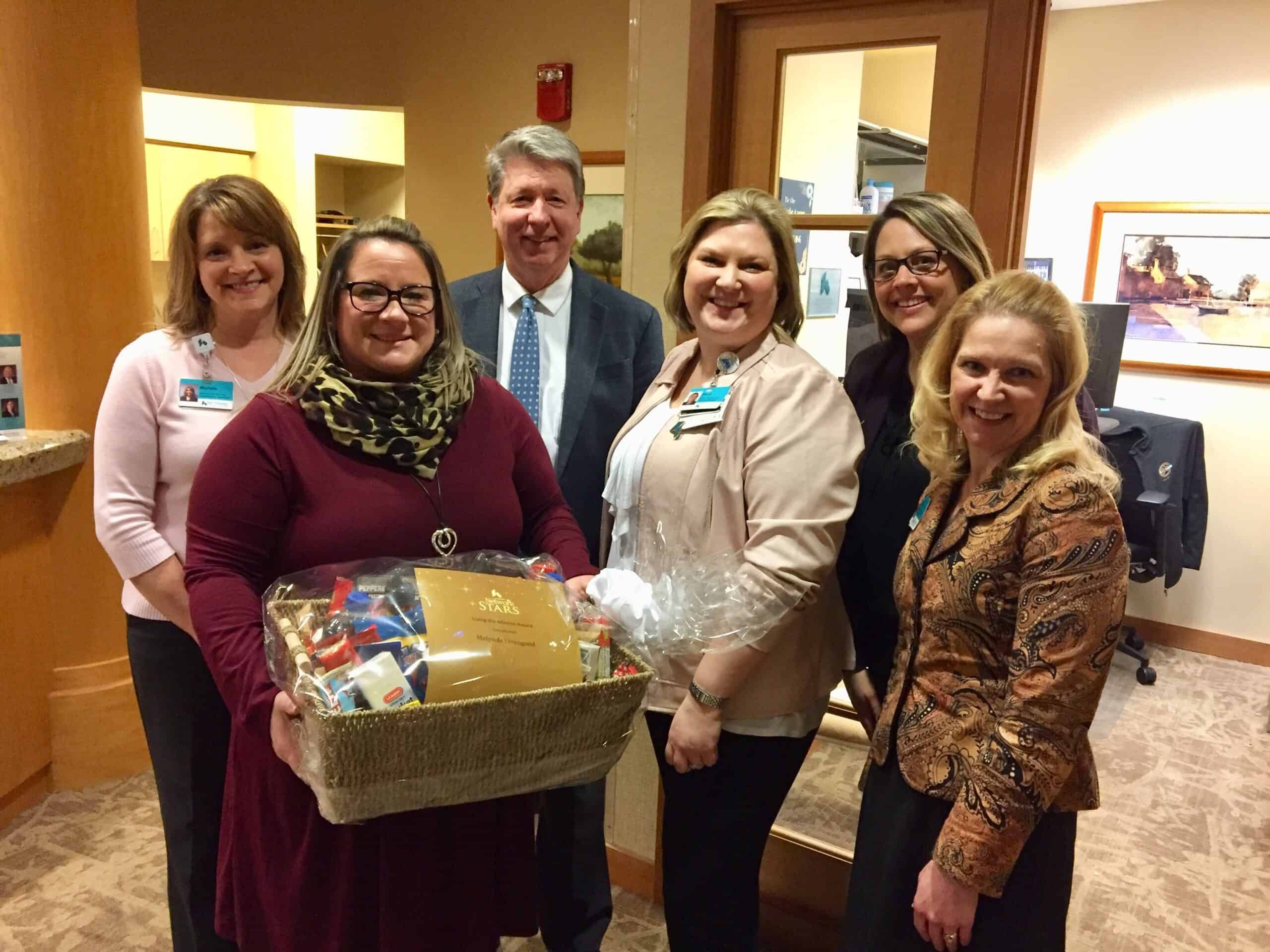 Healthcare team presenting gift basket to woman in burgundy sweater holding award