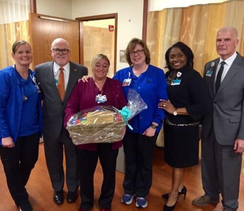 Six healthcare professionals pose together, one holding a gift basket in hospital hallway