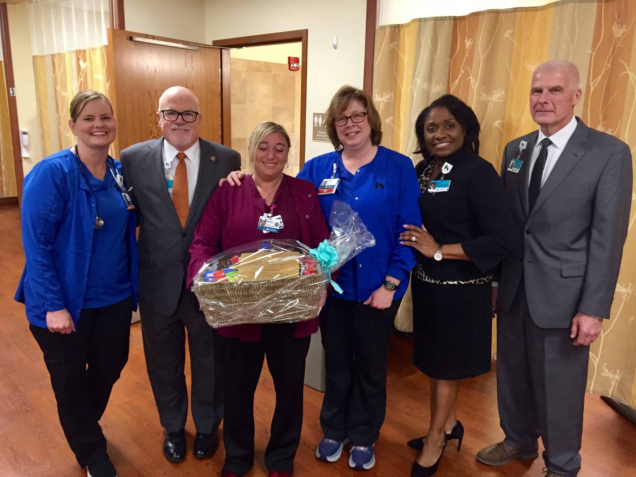 Six healthcare professionals pose together, one holding a gift basket in hospital hallway