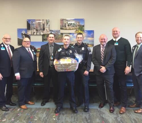 Seven men in business attire and uniforms pose together, one holding a gift basket.