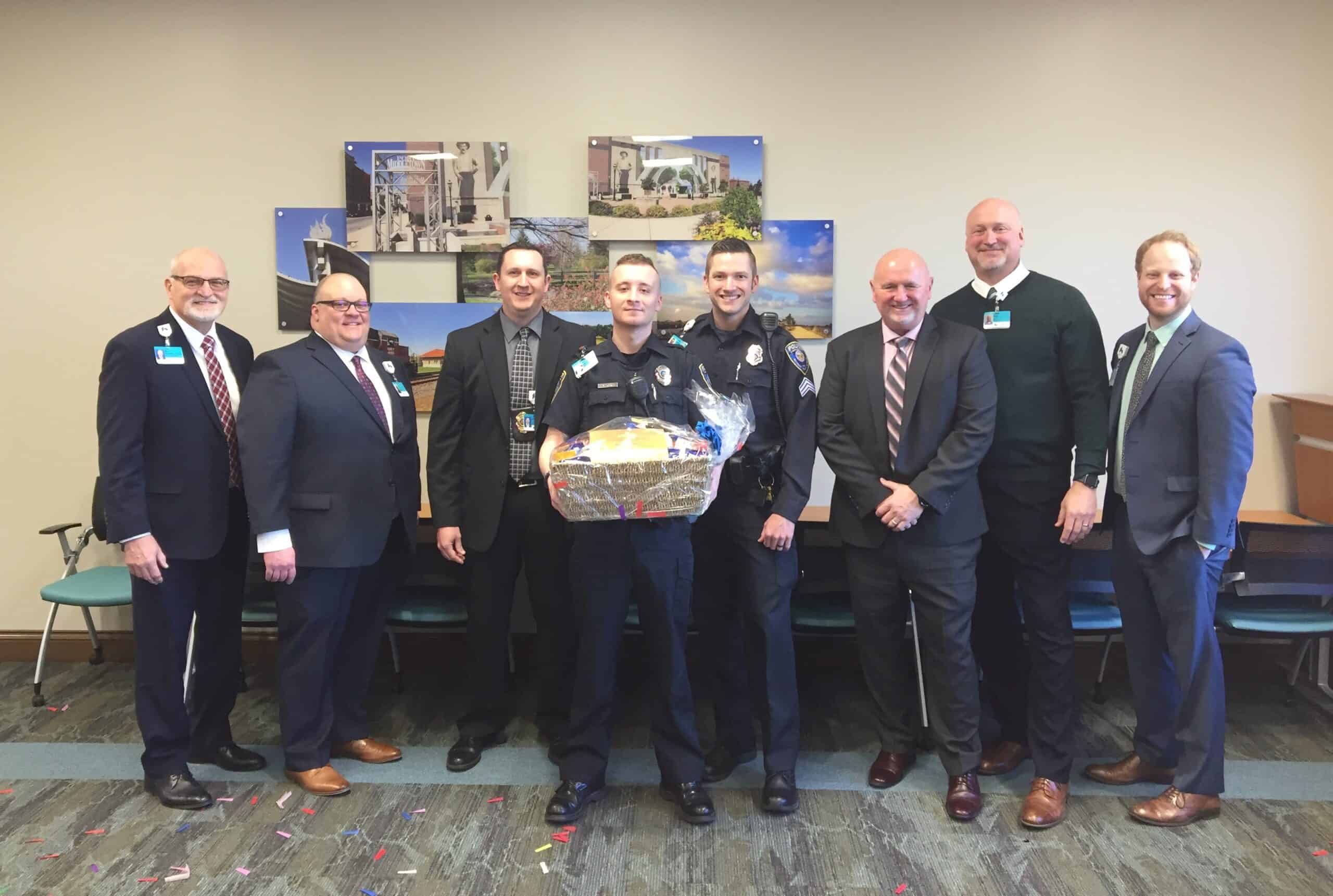 Seven men in business attire and uniforms pose together, one holding a gift basket.