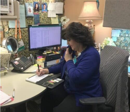 Woman in blue cardigan working at desk with computer and phone in office cubicle