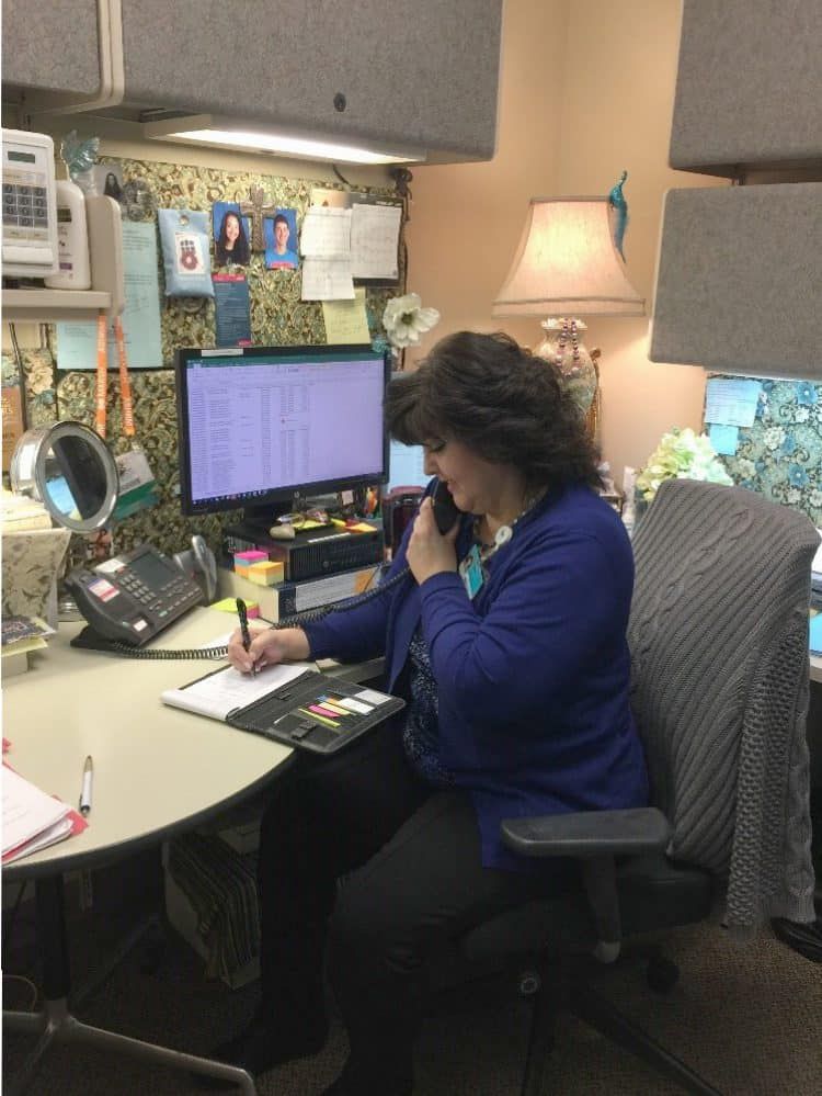 Woman in blue cardigan working at desk with computer and phone in office cubicle