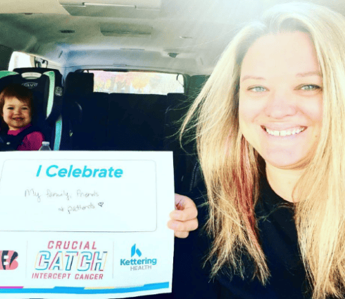 Woman holding "I Celebrate" sign in car with child in backseat, smiling
