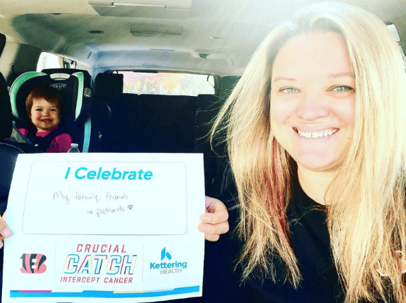 Woman holding "I Celebrate" sign in car with child in backseat, smiling
