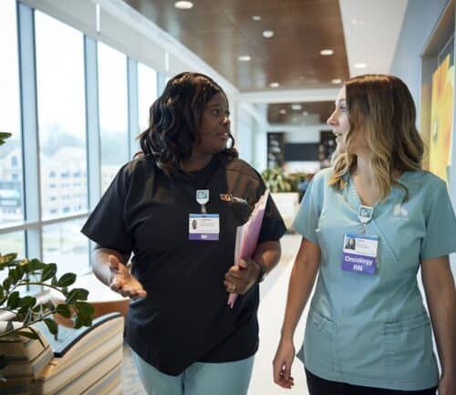 Two nurses in scrubs walking and talking in a modern hospital hallway.