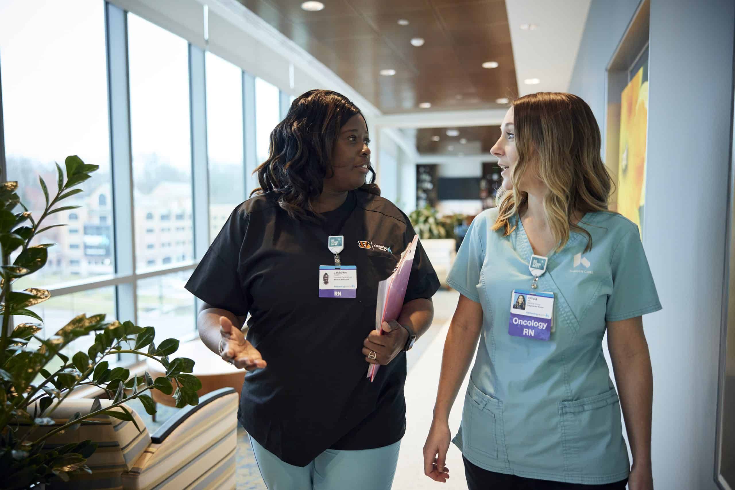 Two nurses in scrubs walking and talking in a modern hospital hallway.