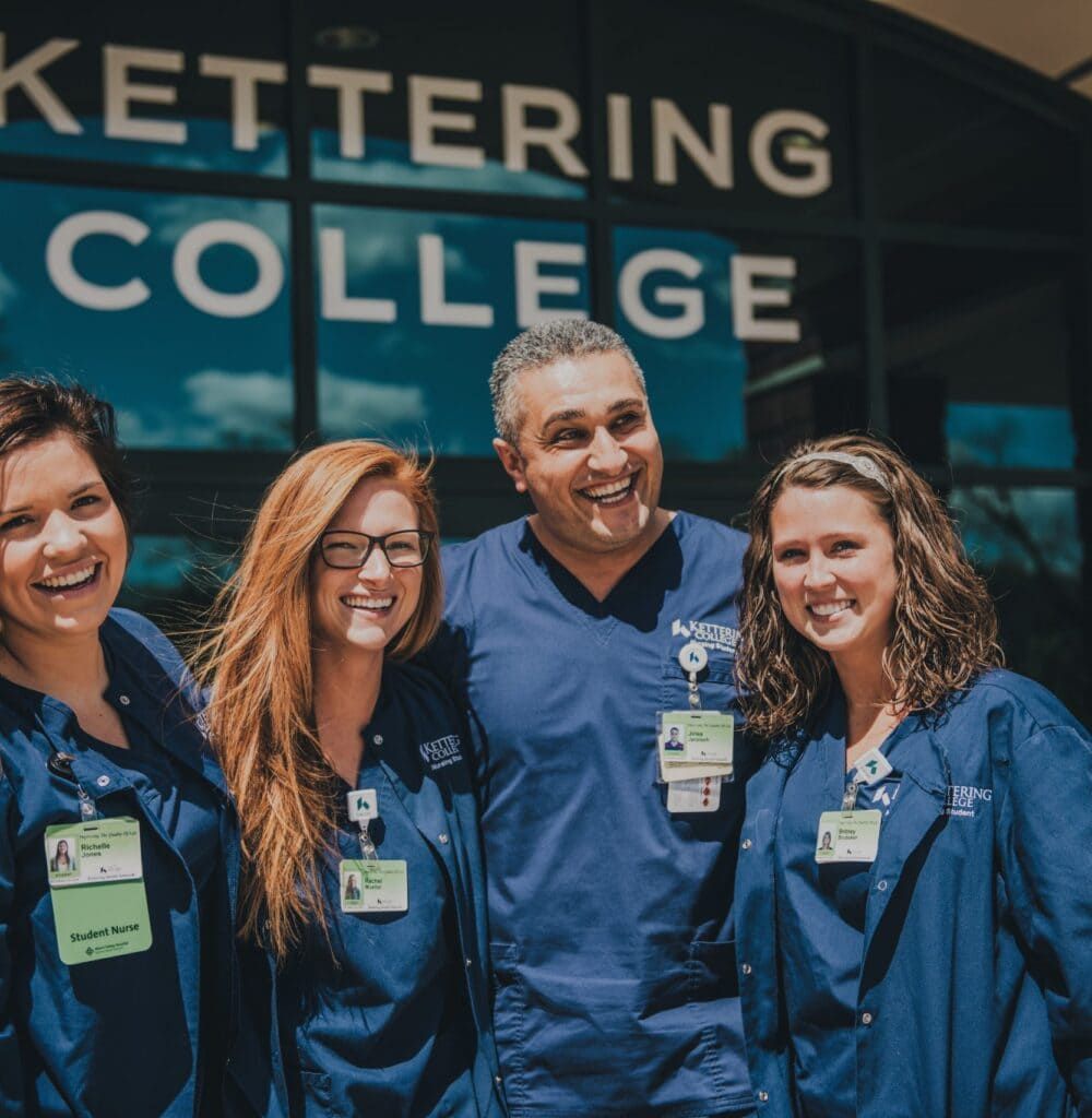 Four nursing students and instructor in blue scrubs smiling outside Kettering College building