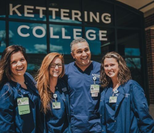 Four nursing students and instructor in blue scrubs smiling outside Kettering College building